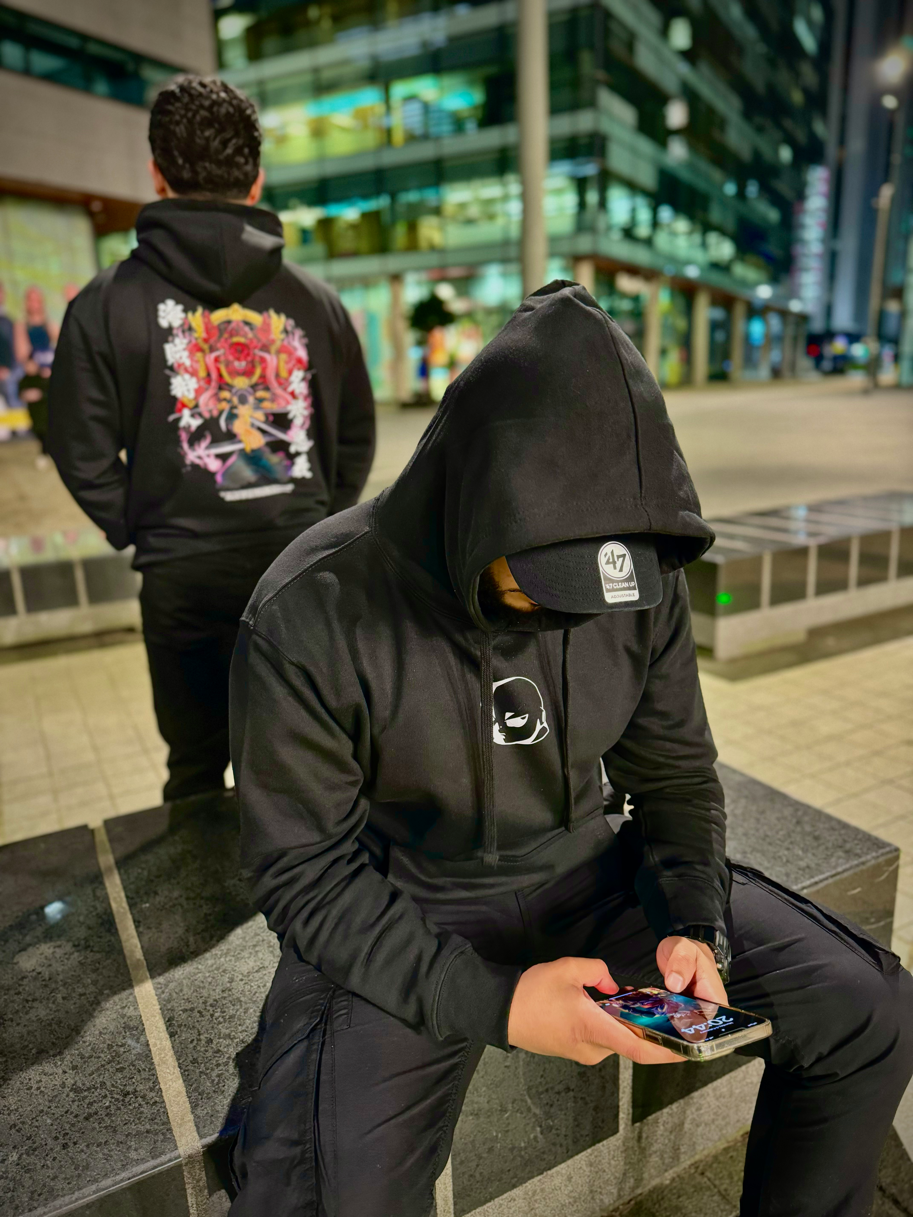 Two male models wearing black Kunai anime hoodies outdoors at night, one seated looking at phone showing front logo, the other standing showing back graphic.