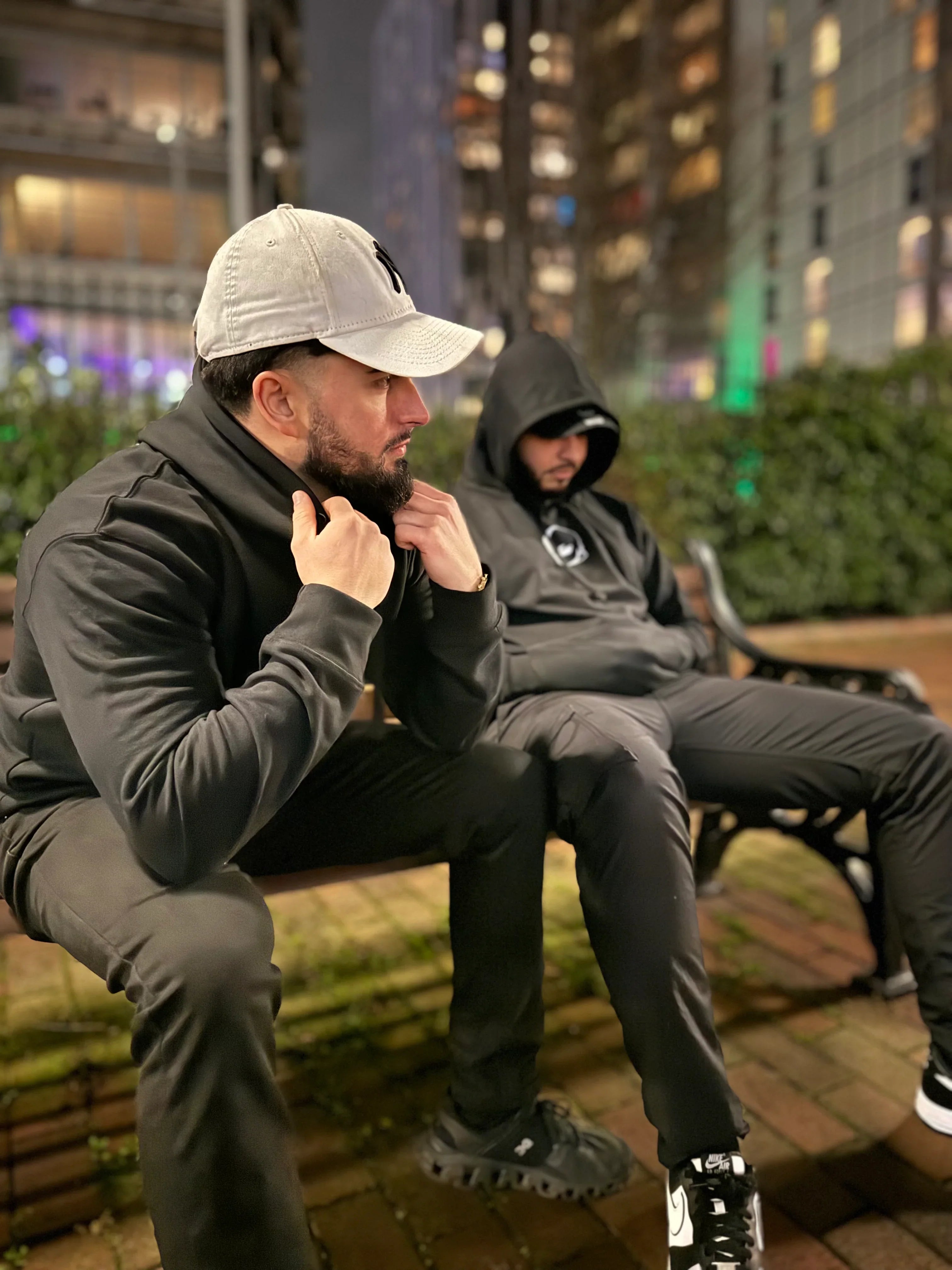 Two men wearing black Kunai anime hoodies while sitting on a bench in a city setting at night, showcasing minimalist anime design.