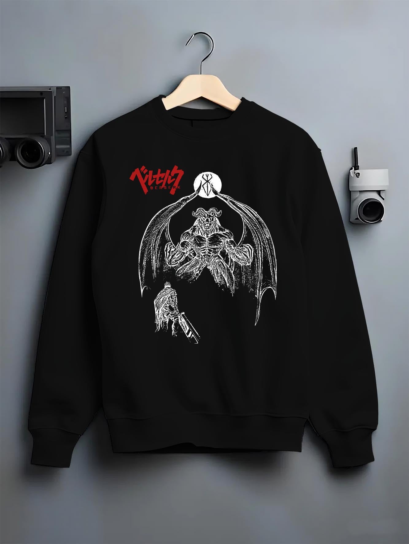 Black crewneck sweatshirt on a hanger against a grey wall, featuring Berserk Guts dark manga panel inspired artwork with apostle demon and gritty retro streetwear style.