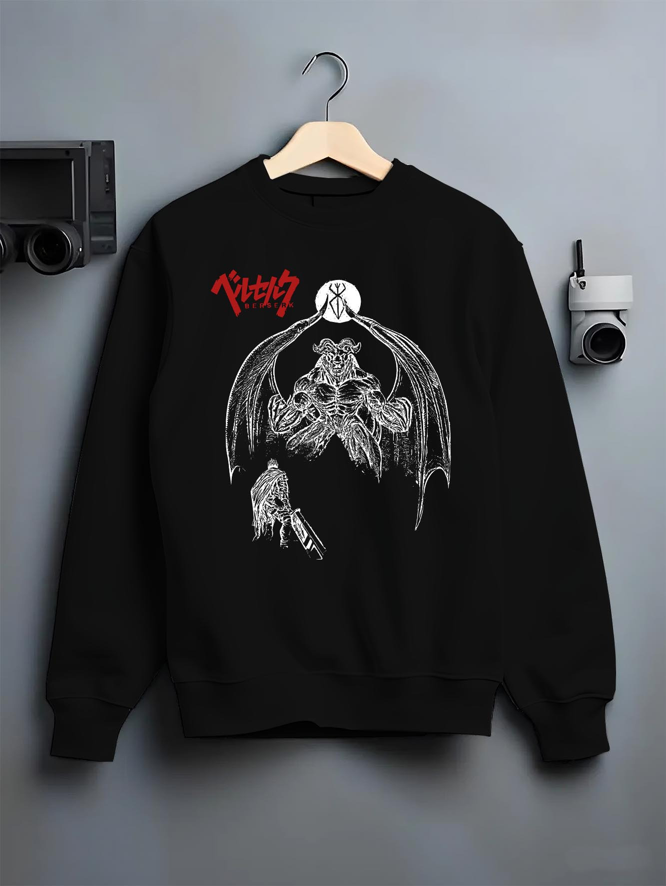 Black crewneck sweatshirt on a hanger against a grey wall, featuring Berserk Guts dark manga panel inspired artwork with apostle demon and gritty retro streetwear style.