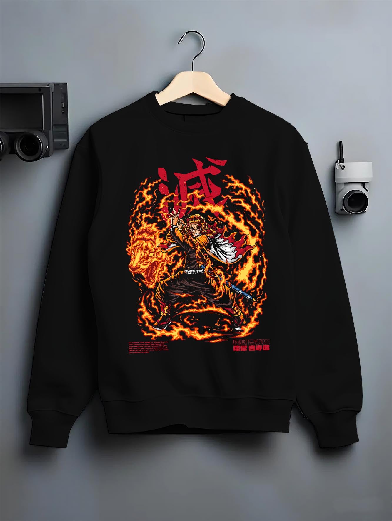 Black crewneck sweatshirt on a hanger against a grey wall, featuring Demon Slayer Rengoku anime poster inspired artwork with bold flame stance and fiery shonen battle style.