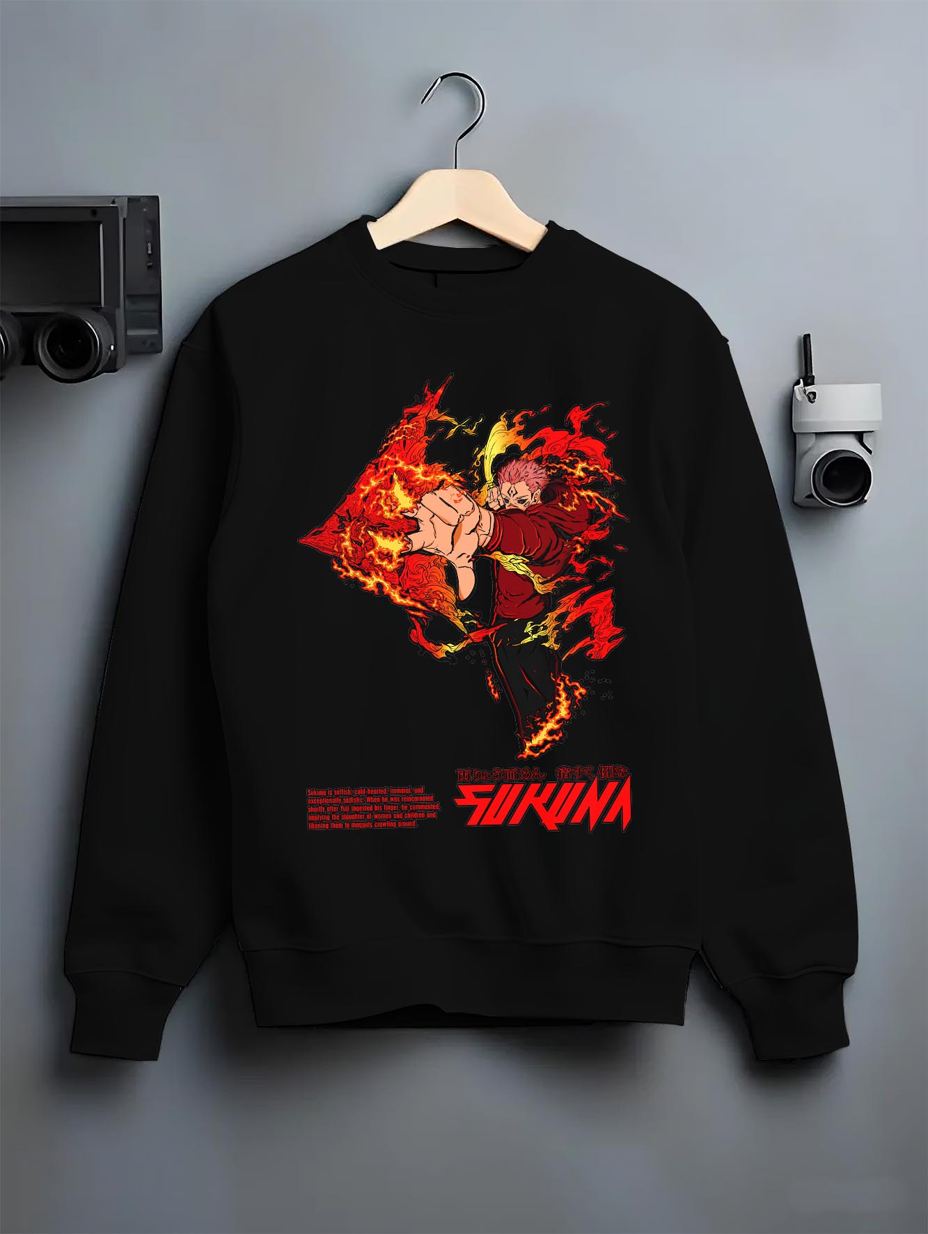 Black crewneck sweatshirt on a hanger against a grey wall, featuring Jujutsu Kaisen Sukuna dark manga panel artwork print with flames and shonen battle streetwear aesthetic.