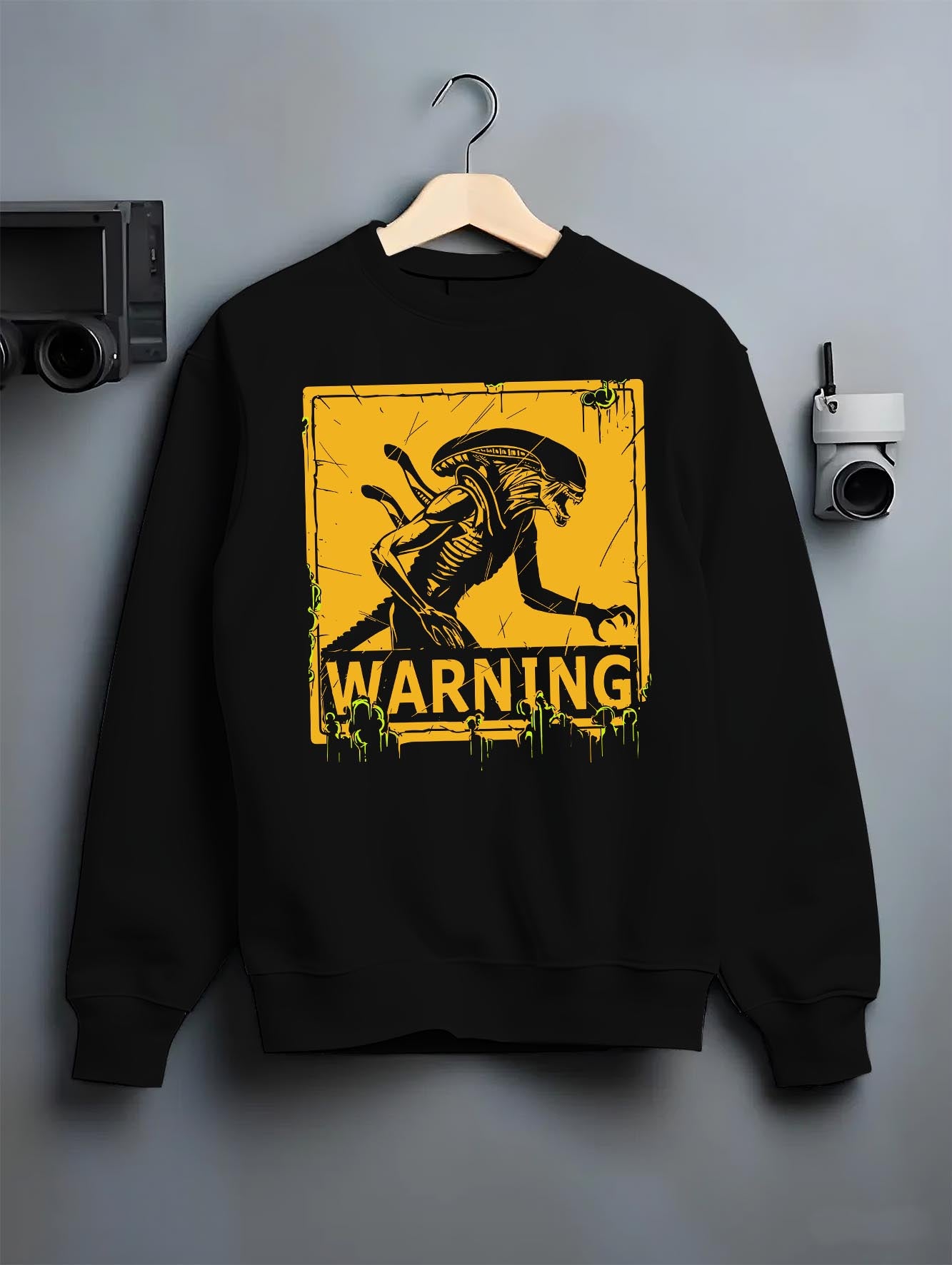 Black crewneck sweatshirt on hanger against grey wall, Alien Xenomorph warning sign graphic with retro sci-fi feel and green drips.