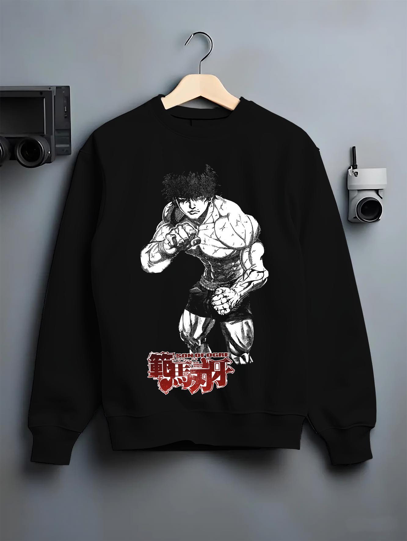 Black crewneck sweatshirt on a hanger against a grey wall, featuring Baki Hanma Son of Ogre anime poster inspired artwork with retro manga style print.