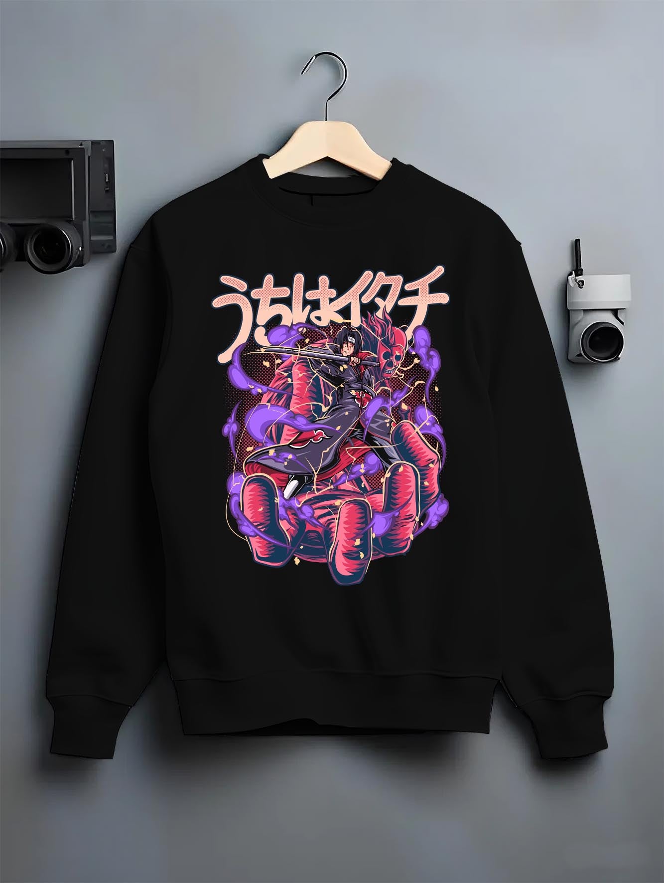 Black crewneck sweatshirt on hanger against grey wall, Itachi Uchiha back graphic with purple Susanoo energy and vintage manga tones.