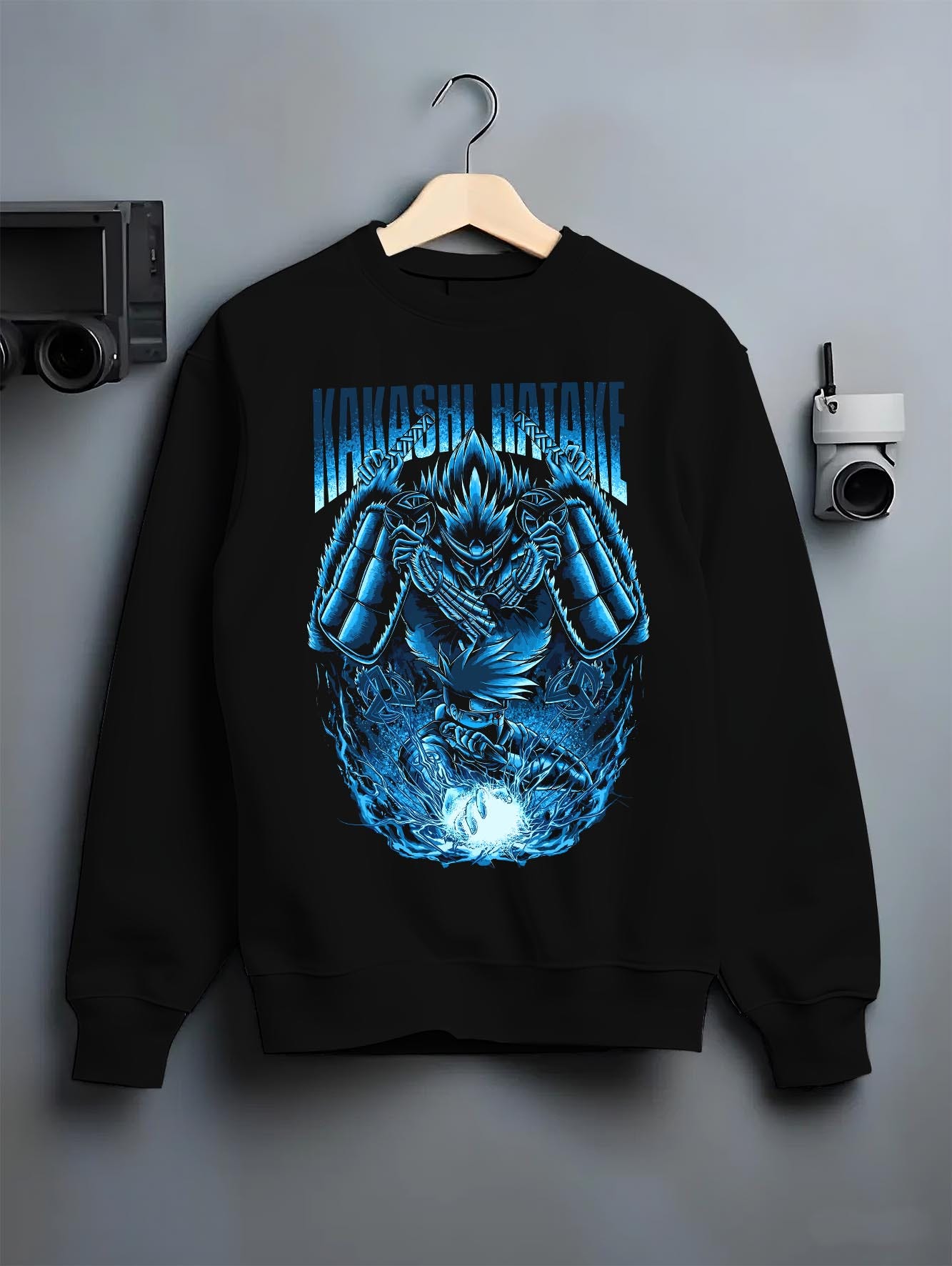 Black crewneck sweatshirt on a hanger against a grey wall, featuring Kakashi Hatake shonen battle character artwork with Susanoo flames and glowing manga detail.