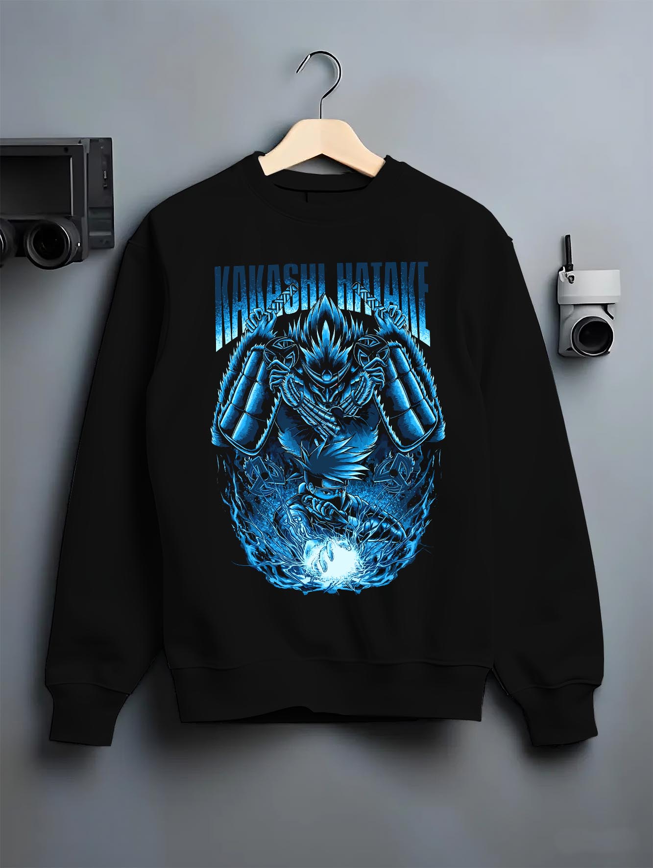 Black crewneck sweatshirt on a hanger against a grey wall, featuring Kakashi Hatake shonen battle character artwork with Susanoo flames and glowing manga detail.
