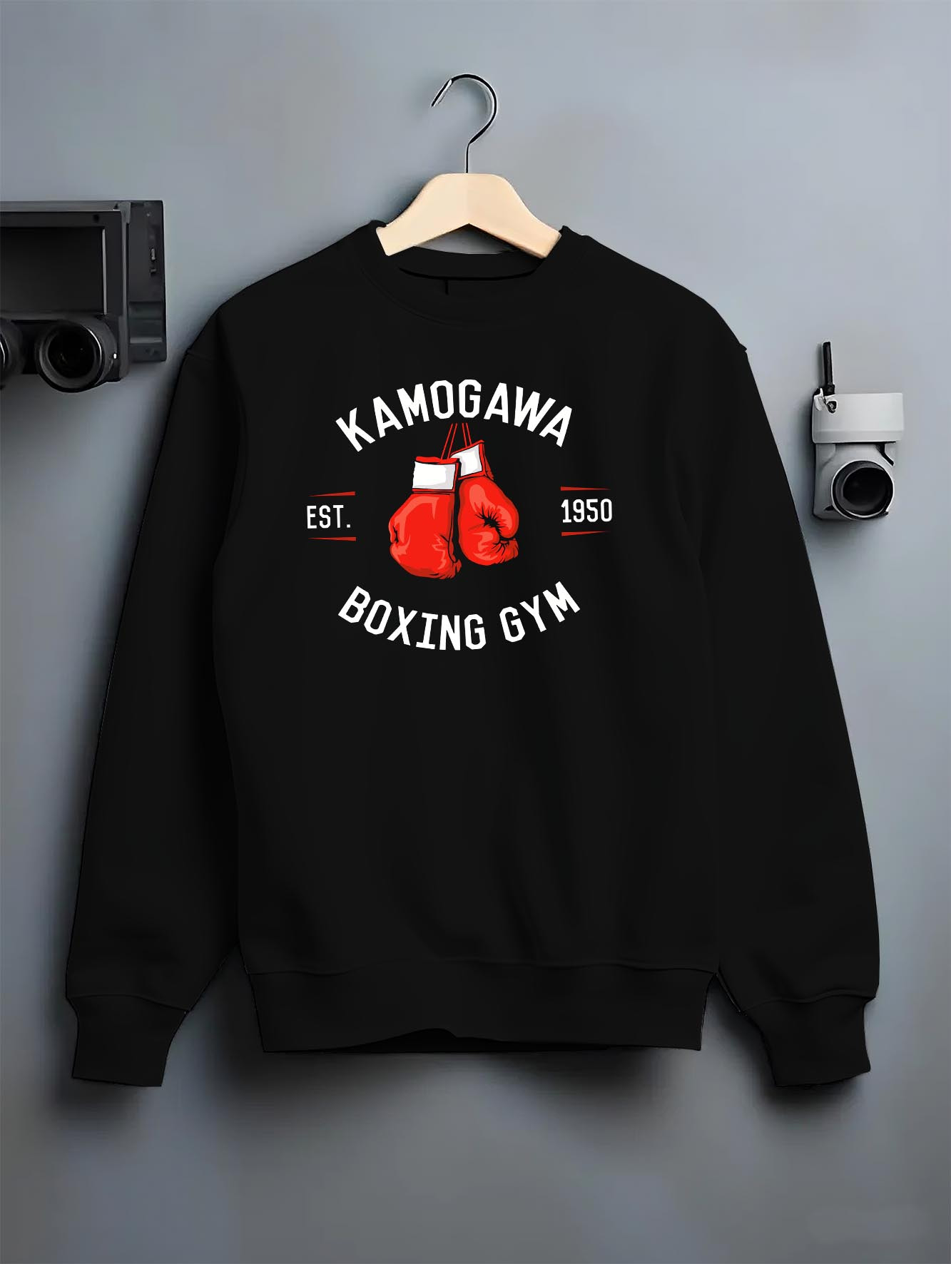 Black crewneck sweatshirt on a hanger against a grey wall, featuring Kamogawa Boxing Gym modern streetwear graphic with vintage boxing gloves and athletic retro vibe.