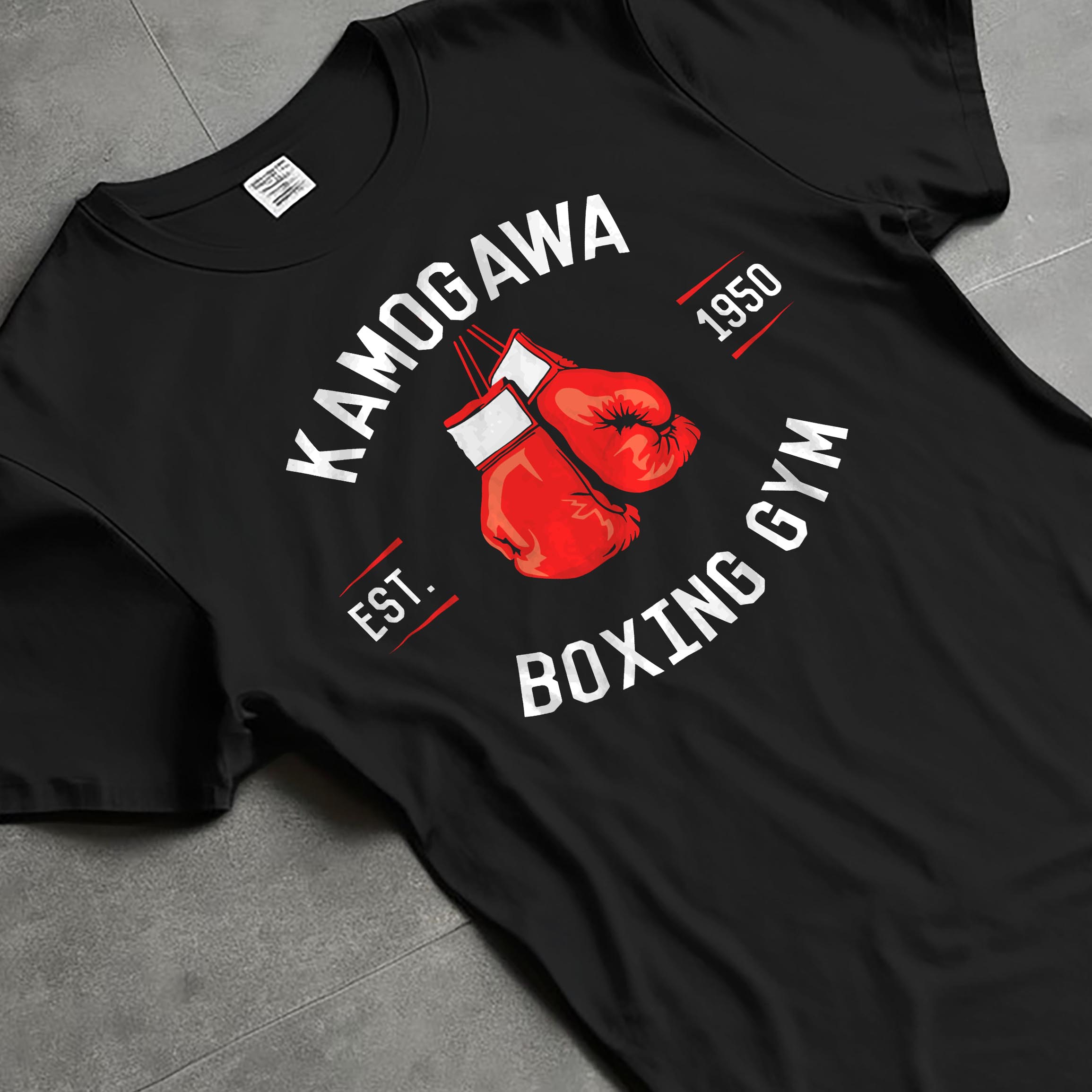 Black T-shirt laid flat with natural fabric folds, featuring Kamogawa Boxing Gym vintage retro aesthetic with boxing glove emblem and bold gym lettering design.