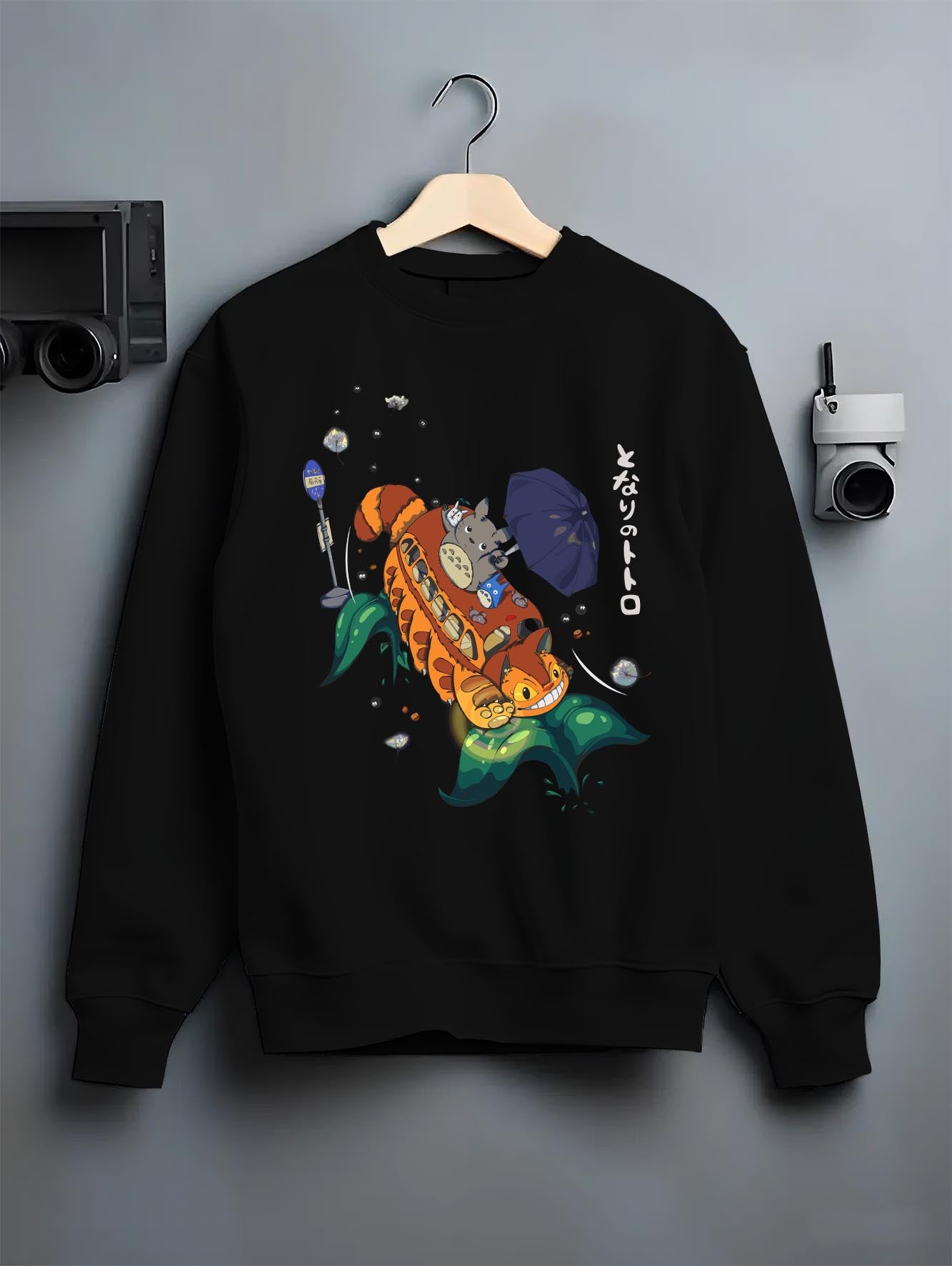 Black crewneck sweatshirt on hanger against grey wall, Totoro with Catbus back graphic featuring umbrella, leaves, and soot sprites.