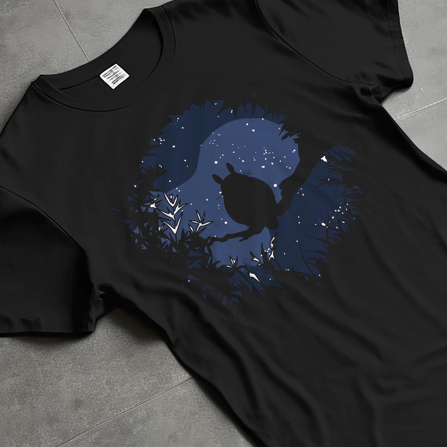 Black T-shirt laid flat with natural fabric folds, featuring My Neighbor Totoro silhouette framed by branches and stars, casual retro graphic.