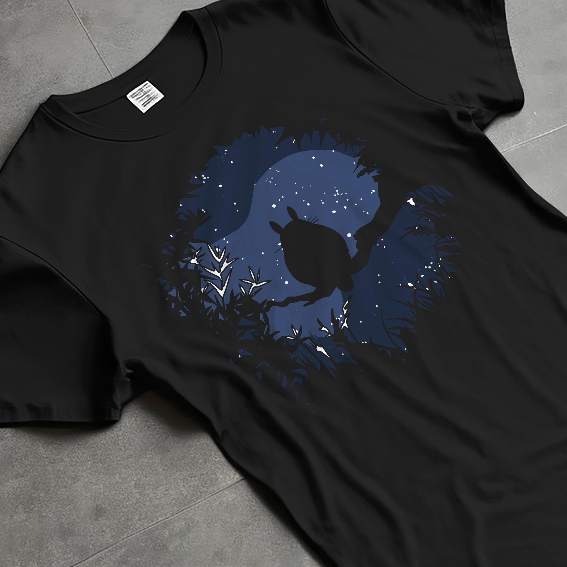 Black T-shirt laid flat with natural fabric folds, featuring My Neighbor Totoro silhouette framed by branches and stars, casual retro graphic.
