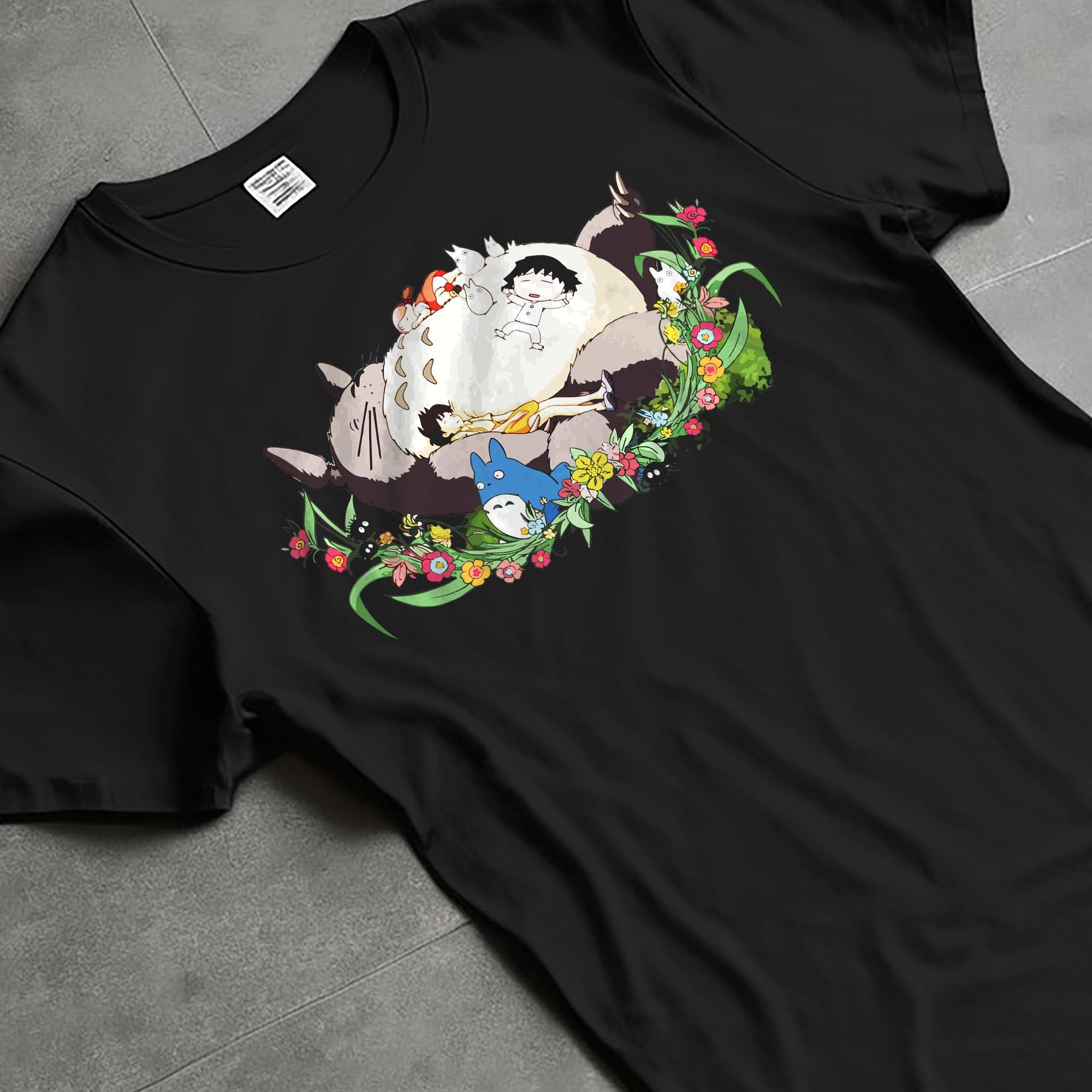 Black T-shirt laid flat with natural fabric folds, featuring Totoro nap tableau and flowers, retro streetwear graphic with vintage anime charm.